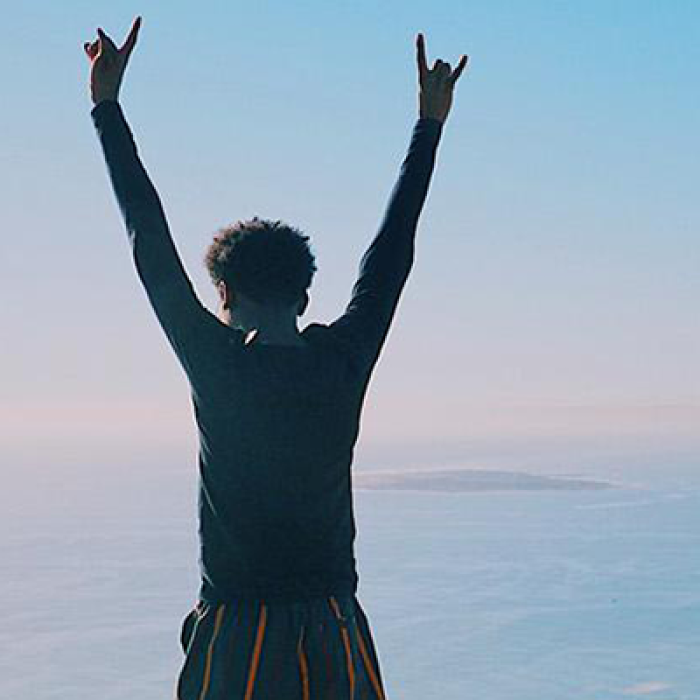 Boy standing on mountain facing horizon, hands reaching towards sky.