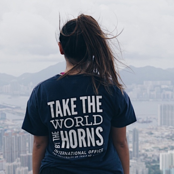 Person standing in front of a hazy city view, with a shirt displaying "Take The World By The Horns".