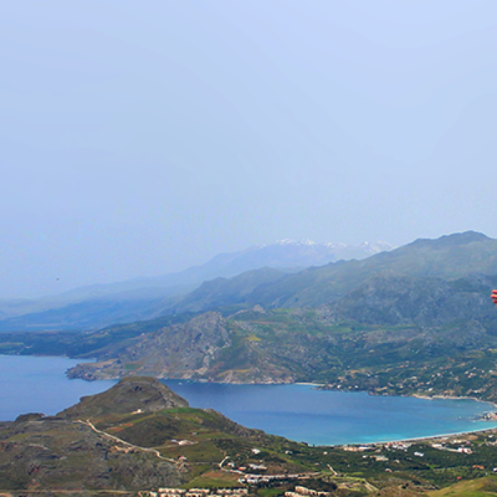 A student stands on a mountain top in Crete, Greece, with arms spread open, overlooking the ocean. Her back faces the camera as she wears a Take the World by the Horns t-shirt.
