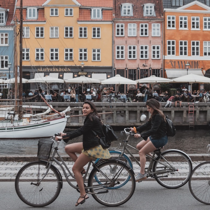 three people riding bicycles near body of water and row of colorful buildings 