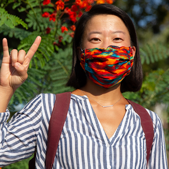 Female wearing blue and white stripped shirt, back-pack, and multi-colored mask making a "hook em" sign with her right hand in front of green foliage with red and orange flowers