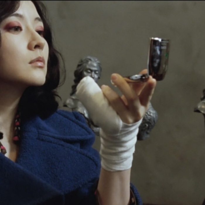 woman in blue blouse holds flip mirror in bandaged hand to look at herself near busts in grey walled room