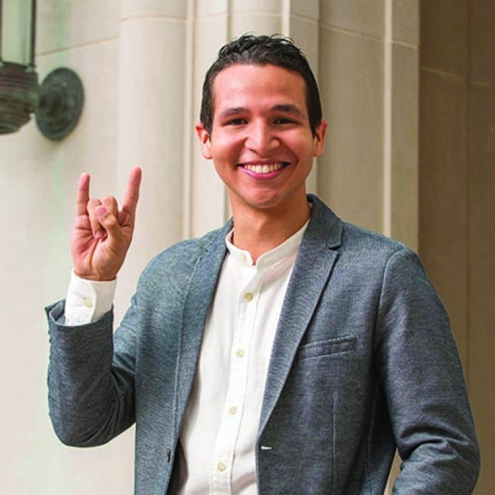 Student in suit doing a Hook'em hand sign in front of UT building wall