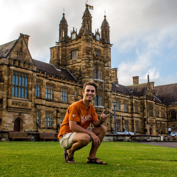 Male student kneeling in front of historic building in Sydney, Australia wearing a UT burnt orange tshirt and giving hook'em hand sign.