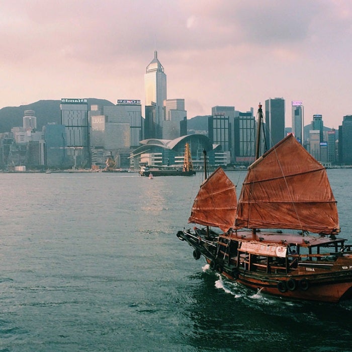 An asian sailboat sails toward the city on calm ocean waters amidst a backdrop of a pink sunset