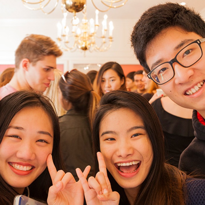 Four international students smiling together in a crowded UT ballroom holding up their hands in a hook 'em sign