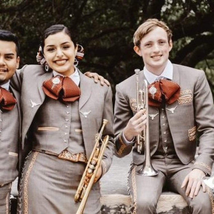 Four UT Mariachi Paredes musicians holding instruments and posing for a group photo wearing grey and burnt orange and gold accented suites  