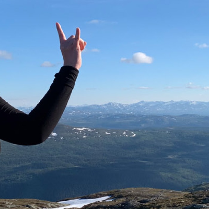 A UT student stands on a mountain on a clear blue sky day with a few clouds overlooking an incredible view while displaying the hookem sign