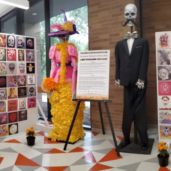 Art Installation Tzompantli Reflexión Artística 2022 showing a female skeleton with a dress make of yellow flowers, pink fabric wearing a purple hat with pink and yellow feathers next to a  male skeleton in a black suit with a bow tie. The skeletons are flanked by installations showing colorful skulls. In the front are marigold plants