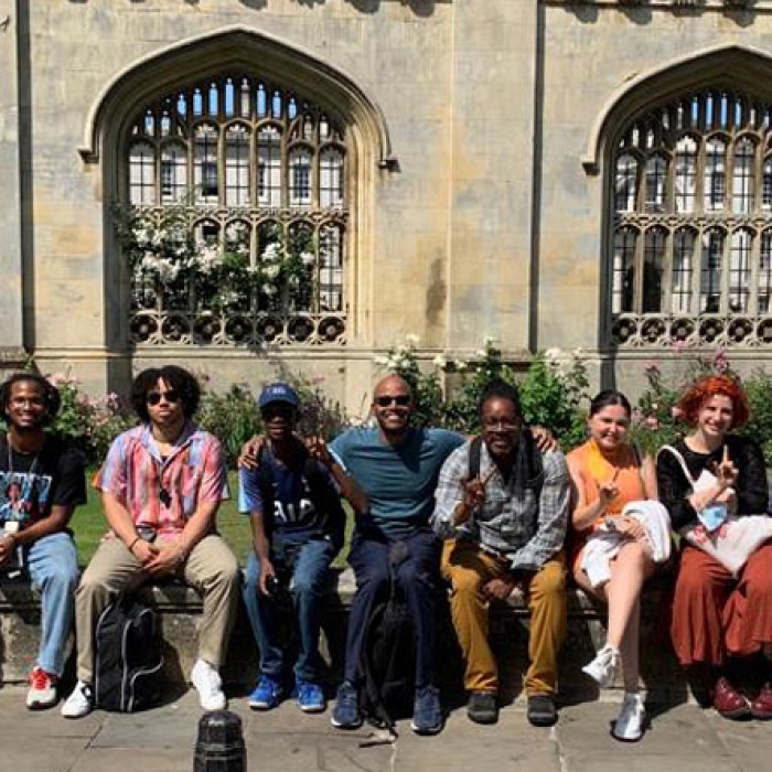 Twelve students and faculty sitting on a ledge in front of Cambridge's Gothic style exterior wall