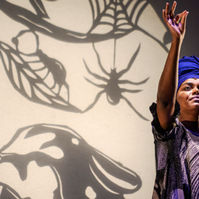 Woman with a blue turban and black robe in on stage looking up towards her index finger while she holds one hand overhead.