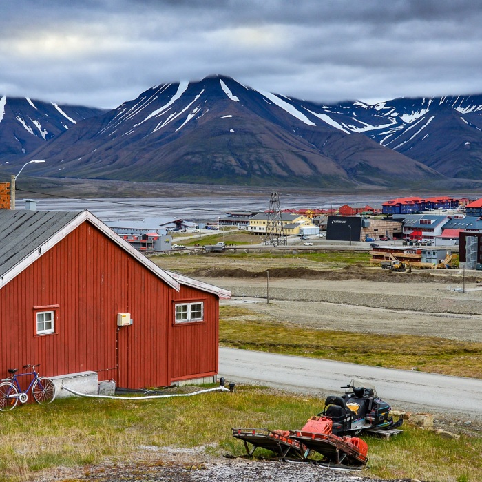 Brick red barns and multi-colored buildings dot the sparse landscape of Svalbard with dramatic snow covered mountain peaks in the background along with an overcast sky