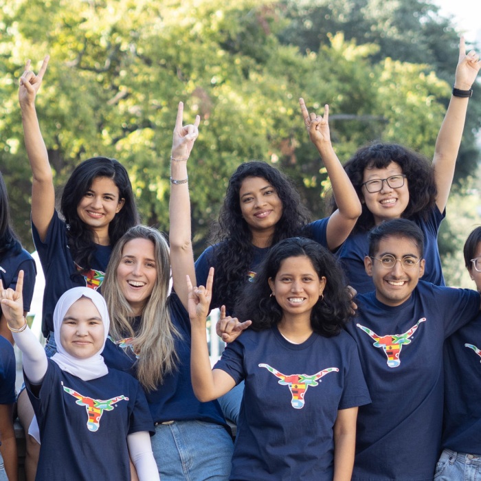A diverse mix of UT Students show off their smiles and longhorn pride in this group shot with live oaks and campus buildings in the background