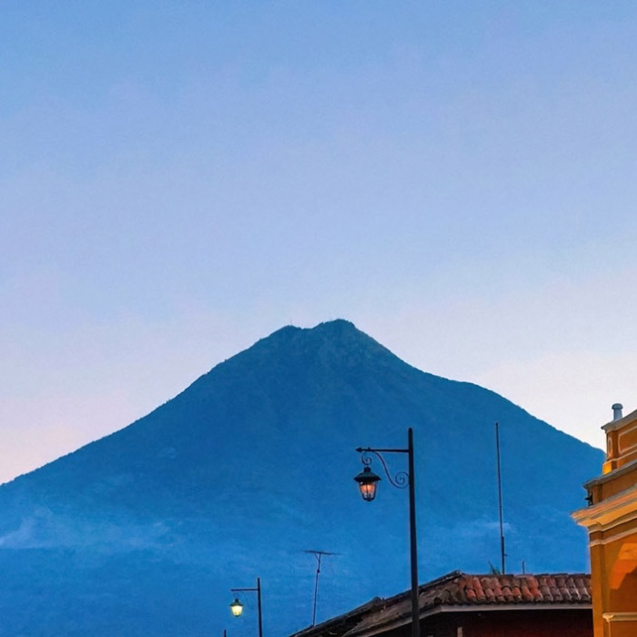 A perspective view of an old stone street and historic buildings lit up in the foreground framing the distant Agua volcano in Antigua, Guatemala during the blue hour of the evening.