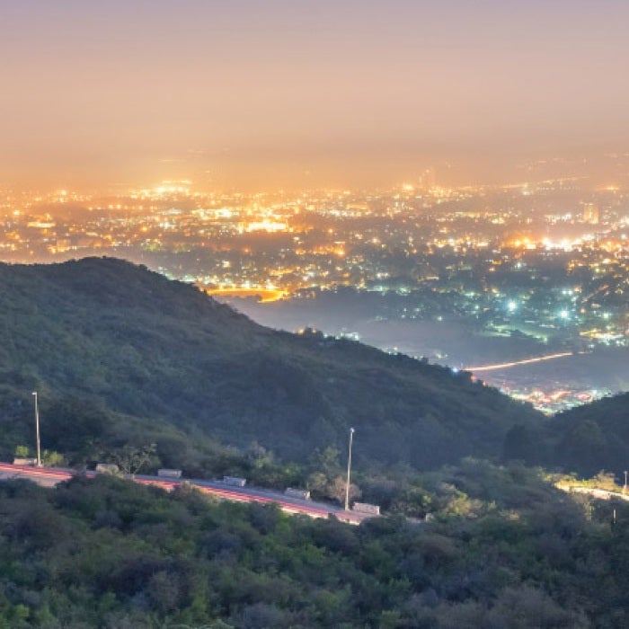 A highway road in a mountain overlooks a city of lights at dusk above a hazy city-sky.