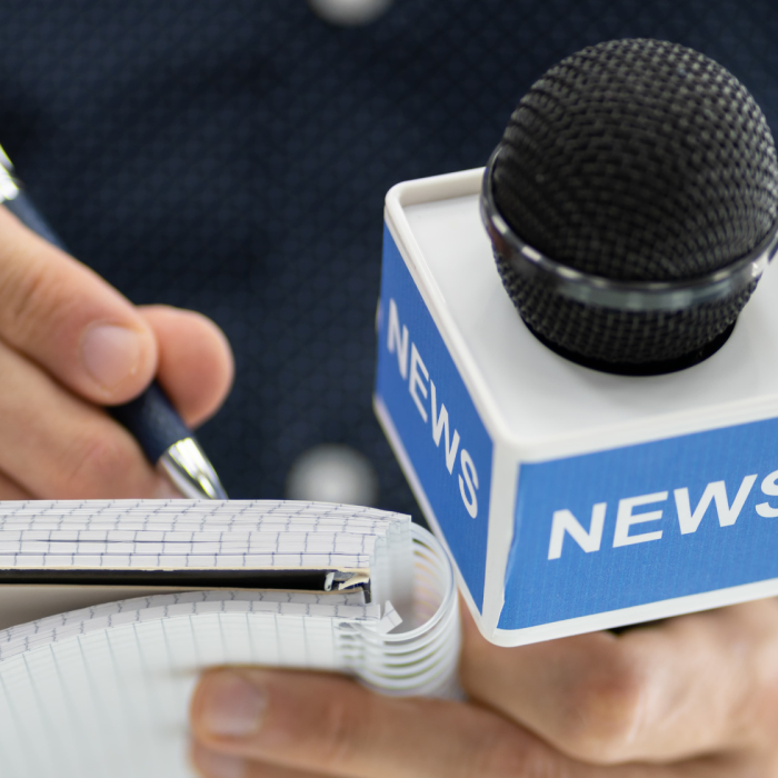 close up image of two hands holding a "news" microphone and writing notes on a small notepad