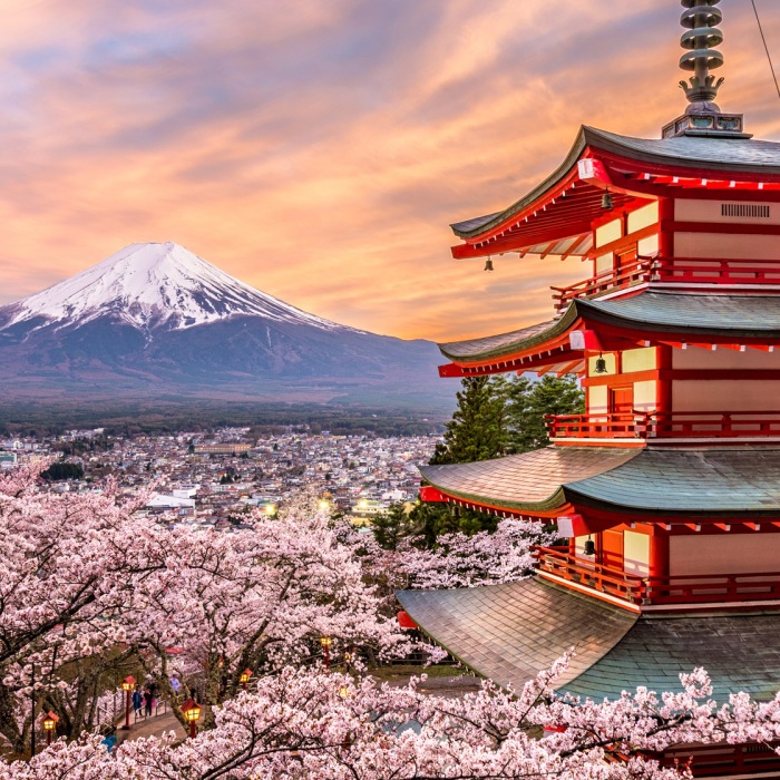 The bright red and green roofed Chureito Pagoda stands proudly amidst walkways lined with cherry blossoms with the majestic Mt. Fuji, backdropped by a pink and orange sky
