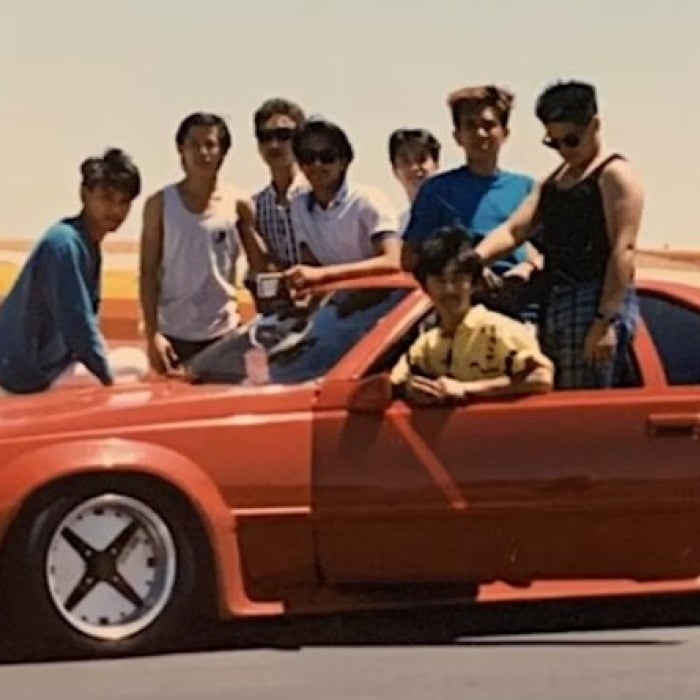 A group of men gather around and on top of a red vintage car. 