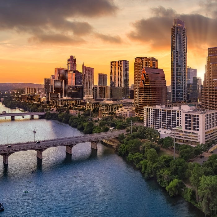 An aerial view of Lady Bird Lake and downtown Austin. You can see the Congress, South First, and Lamar Bridges. You can see the sunsetting behind the buildings.