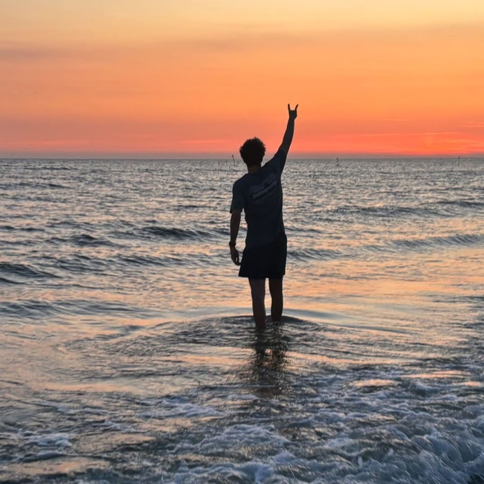 A young man displays the Longhorn hook'em while standing in the surf during a colorful sunset