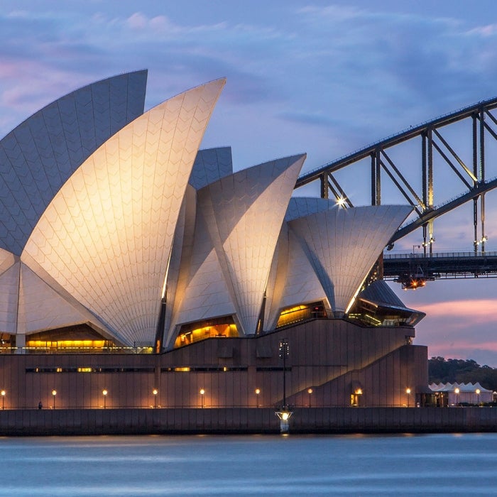 The Sydney Opera House in Australia is illuminated and backdropped by the bridge beside and beautiful pink and blue sunset during the blue hour of the evening