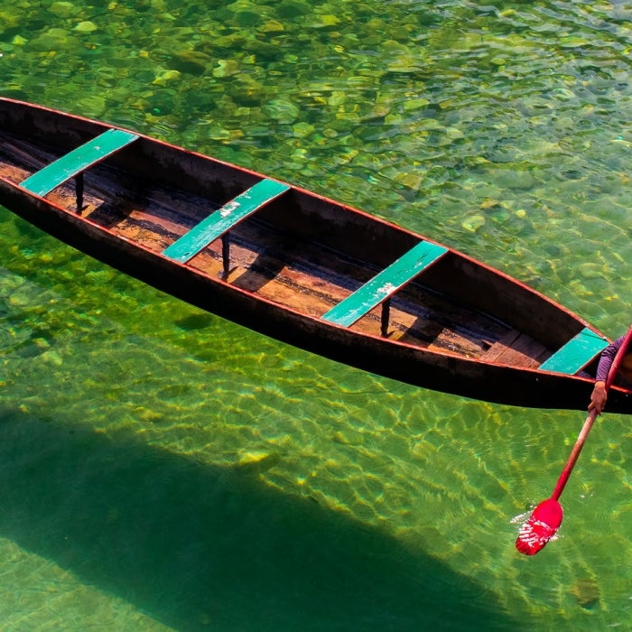 A young man sits on the end of a wooden boat with red paddle over clear green water