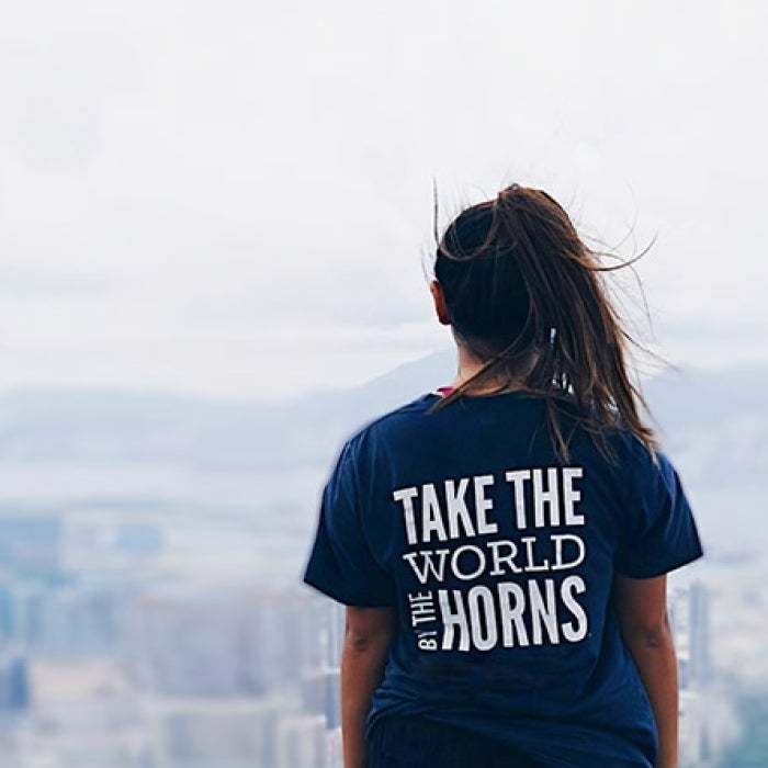 Female looking out on city landscape with blue t-shirt reading Take the World by the Horns