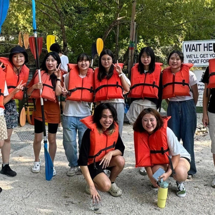 Smiling ELC students wearing lifejackets with paddles and kayaks pose in front of Barton Springs