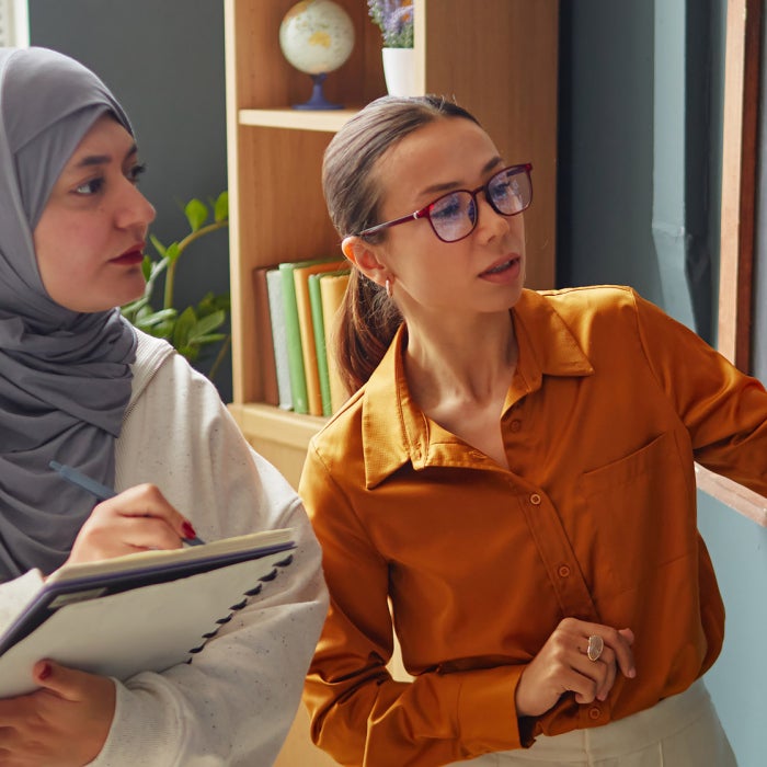 A female professor points at cursive english writing on a chalk board as a foreign teacher wearing a hijab takes notes