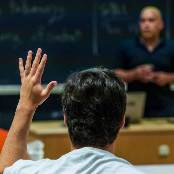Students raise their hand with questions for a professor in the background