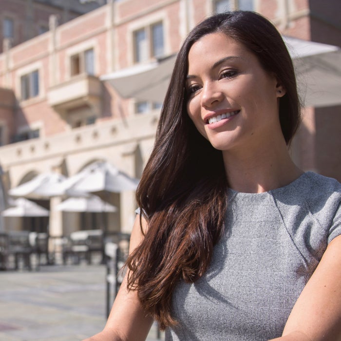 A young smiling women with long black hair uses a laptop at an outdoor courtyard in a European country