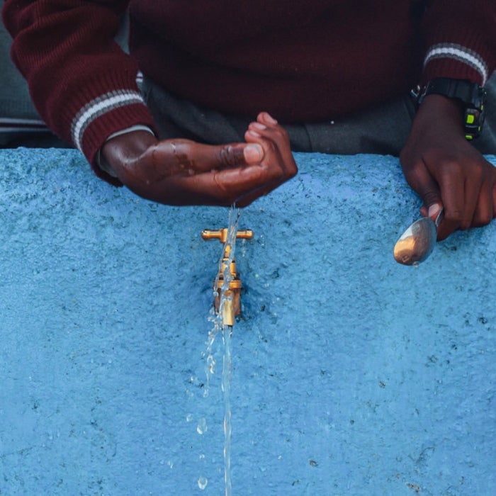 Tanzanian hands scooping and drinking water from brass fountains set against a light blue stone wall