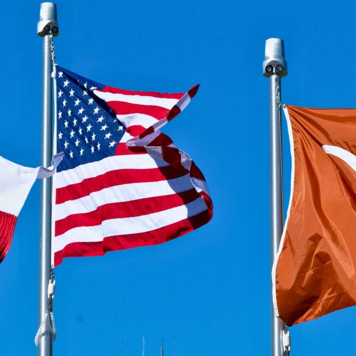 Texas state flag, American flag, and University of Texas Longhorns flag waving against a clear blue sky.
