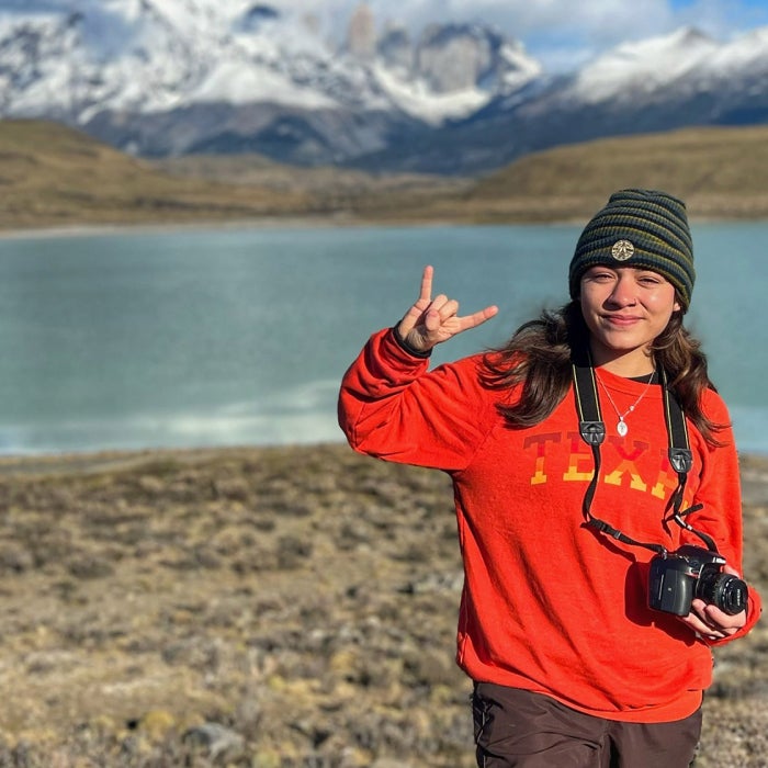 A female student wearing a read Texas sweatshirt holding a camera and posing with UT hook'em sign in front of an alpine lake