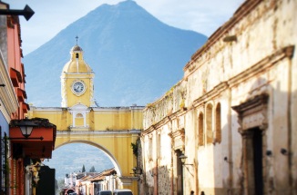 Rows of colorful buildings line the street leading to the famous yellow Saint Catalina archway in historic Antigua Guatemala with Agua volcano looming above in the distance.