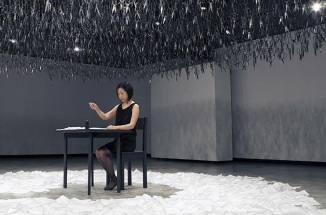 woman sits at table in large room with hanging art installation above and white petals on ground
