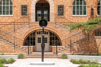 Sentinel IV, a bronze sculpture with a thin, long body with a large half-sphere head, stands in the middle of the Anna Hiss Gymnasium Courtyard on a bright day.