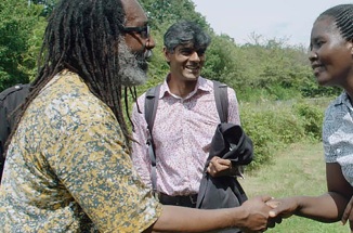 Raj Patel watches while Anita Chitaya and a man shake hands in a field