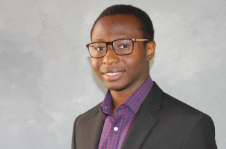 A researcher from Nigeria in a suit poses for a portrait in front of a gray backdrop.