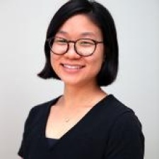 Lena Suk in black top with round black-framed glasses, smiling into camera. Headshot.