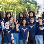 A diverse group of UT students smile while wearing blue Texas Global shirts and displaying the hook'em sign