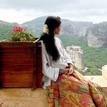 A female abroad student with a white shirt and colorful skirt sits in an ancient balcony that looks out to a valley filled with trees and towering mountains in the background