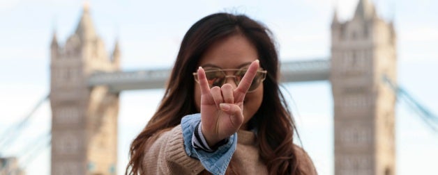brown haired woman wearing glasses holds up hand gesture with pinky and index finger pointed outwards in front of face with two towers and bridge in background