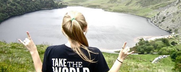 girl with pony tail faces away from camera throwing up hand signs near green valley and body of water during daytime