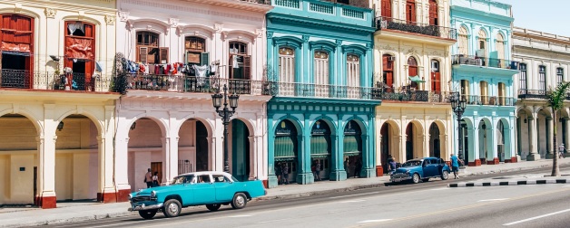 two vintage cars parked outside row of colorful buildings 
