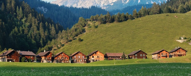 row of brown houses in green hills near trees and snowy mountains during daytime