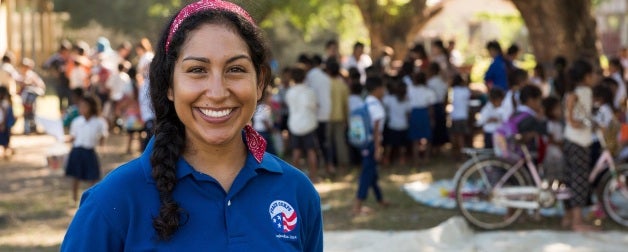 a woman peace corps volunteer smiles, wearing a peace corps shirt 