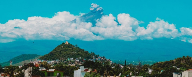 Aerial view of mountains with city of Puebla below with blue skies against tall volcano backdrop that rises above the clouds