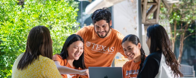 A group of diverse students gather around a laptop at the UT campus on a sunny day