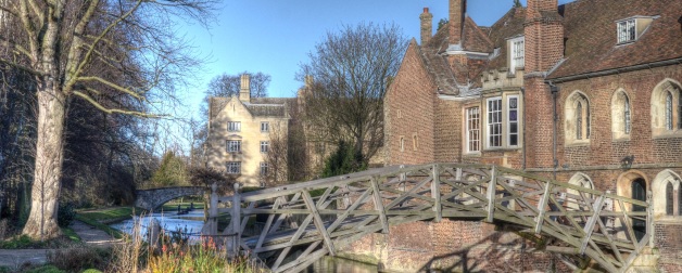 alt=creek running through rural England with a wooden bridge across it and brick cottage to the right